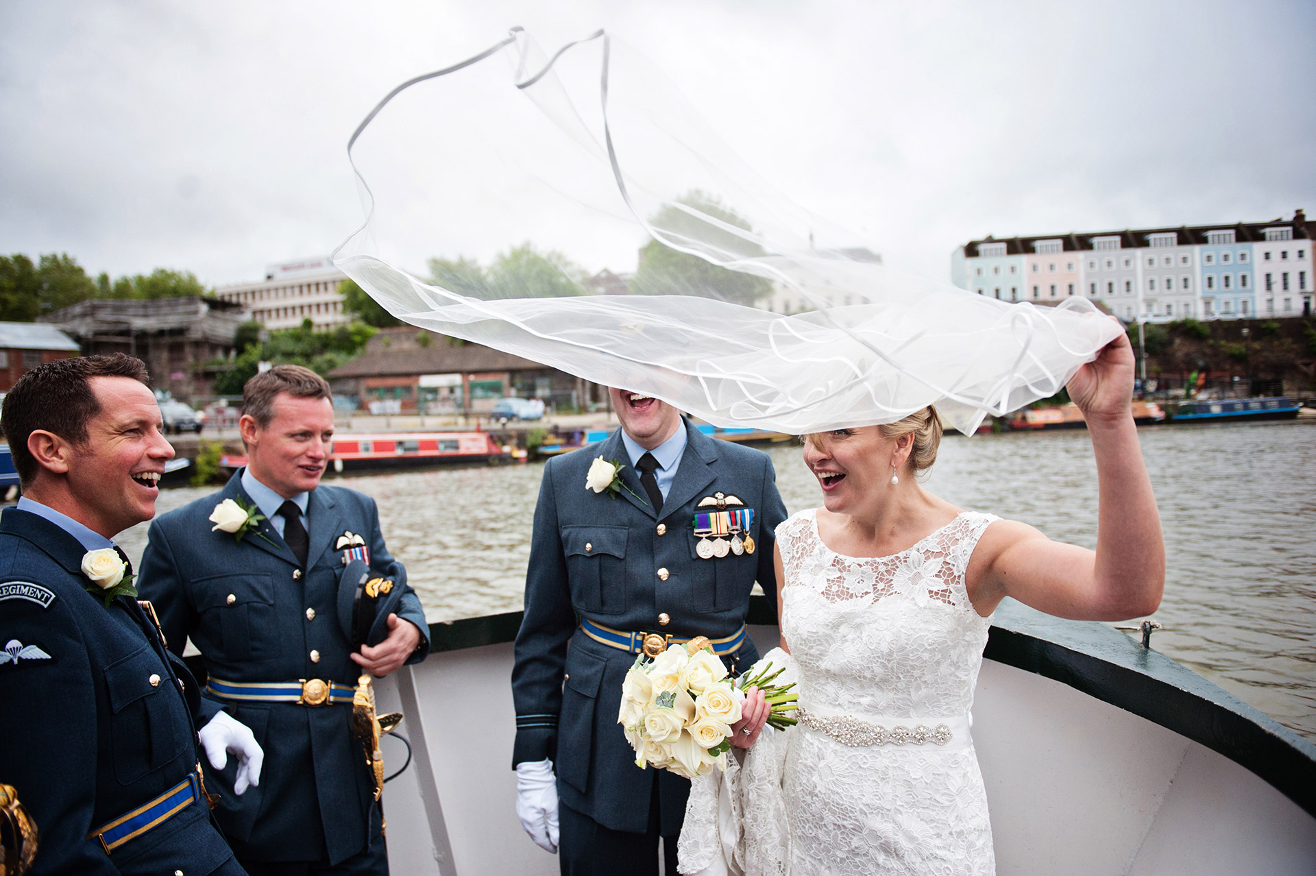 Bride with her veil being caught by the wind. Bride and groom and groomsmen on a boat in Bristol. Groomsmen wearing UK military uniform. Everyone laughing.