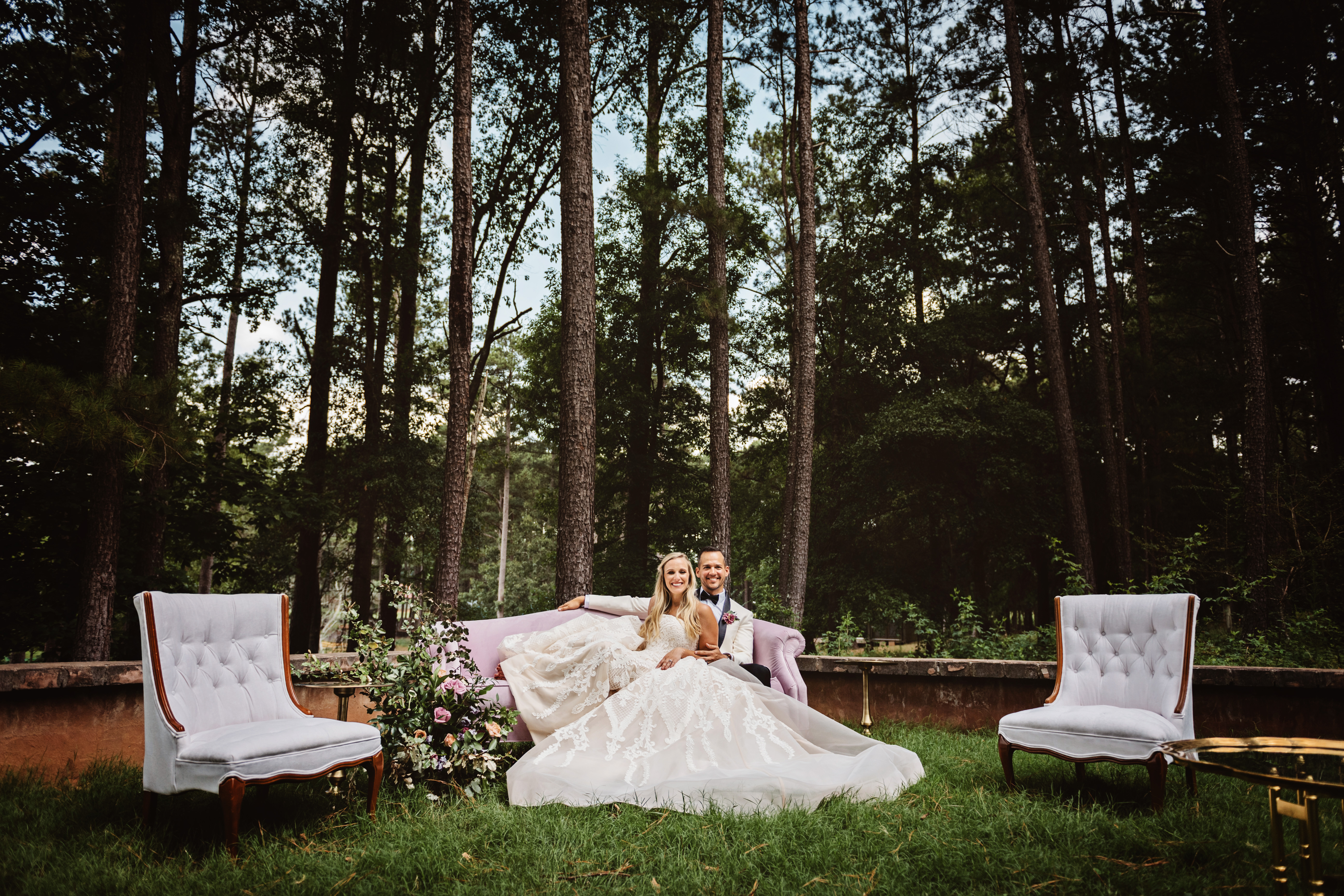 bride and groom sit back on a glamorous sofa smiling, in the woods in Atlanta, Georgia
