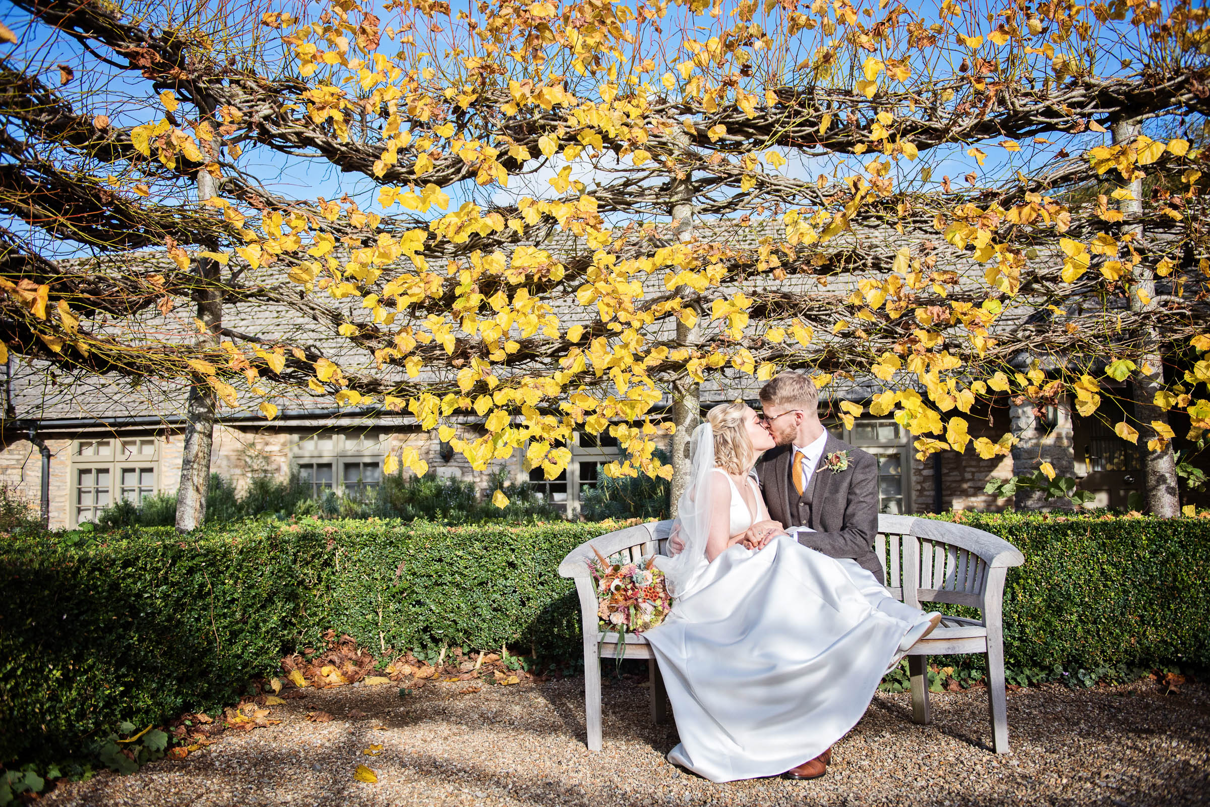 Bride & Groom sit on a bench surrounded by autumnal colour having a quick kiss!  