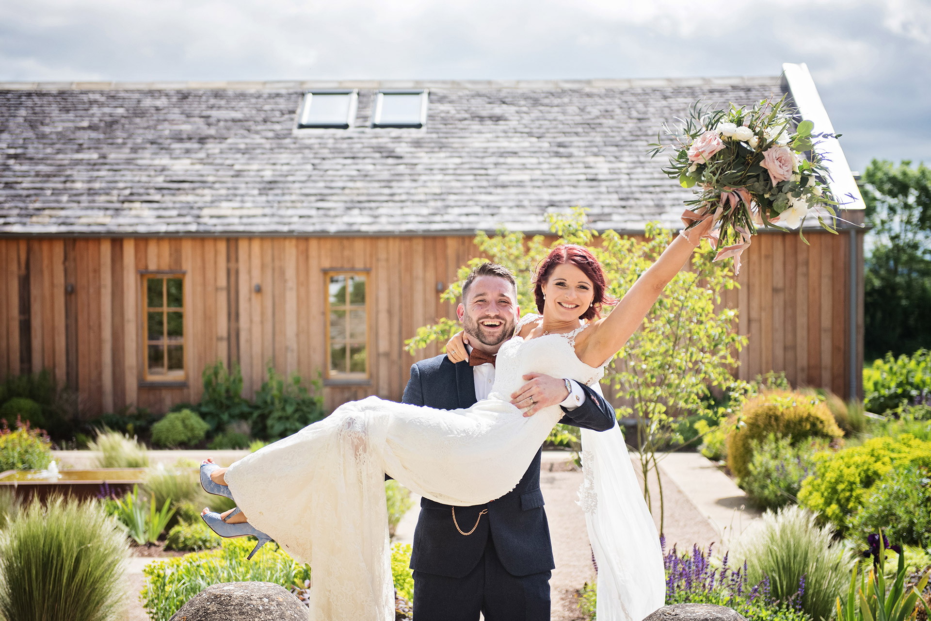 Groom carrying his new bride, with her arm raised holding her bouquet. Wooded barn and garden in the background.   