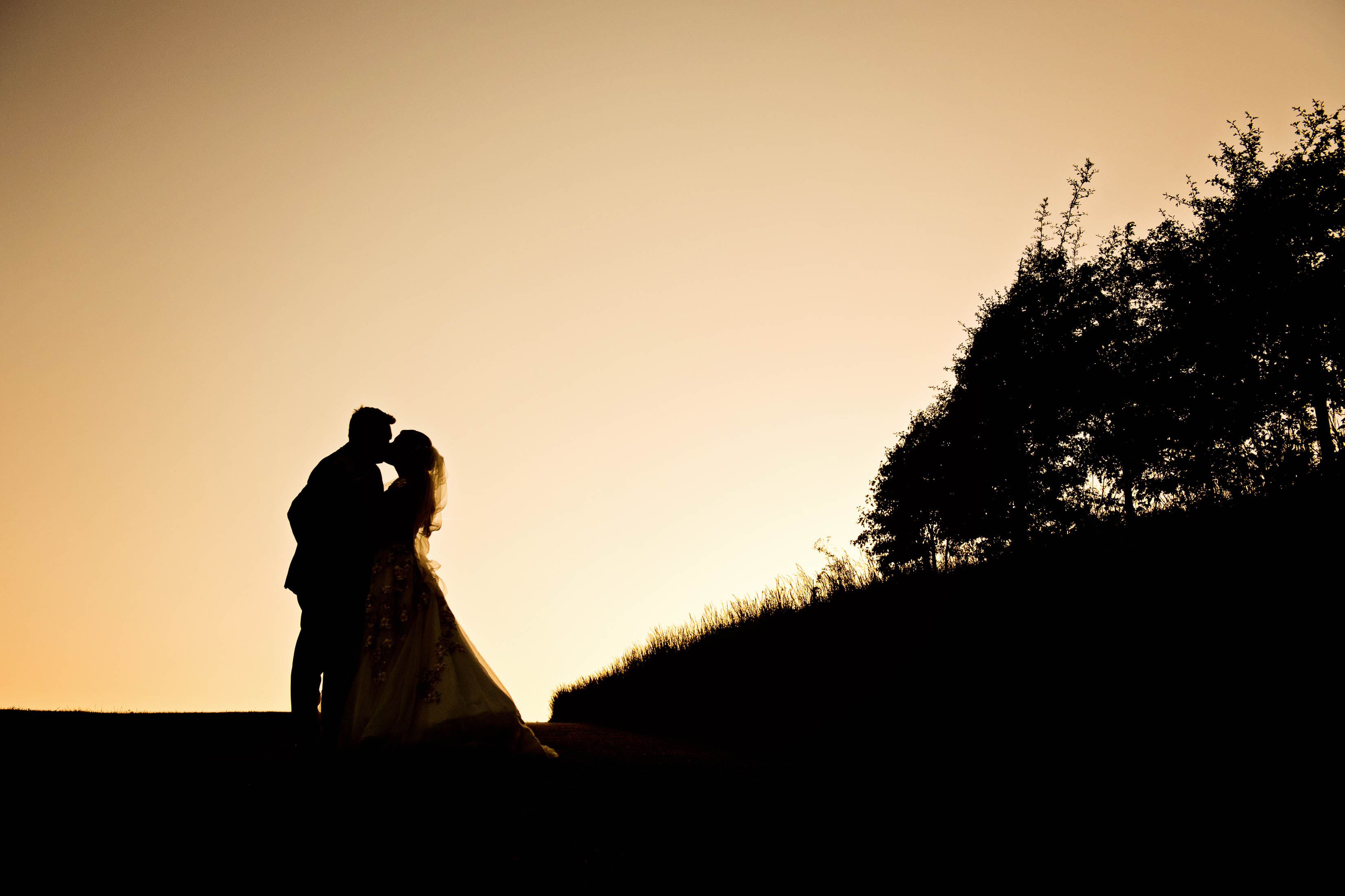 Bride and groom kiss, silhouetted from the sunset behind them. Taken at The Kingscote Barn