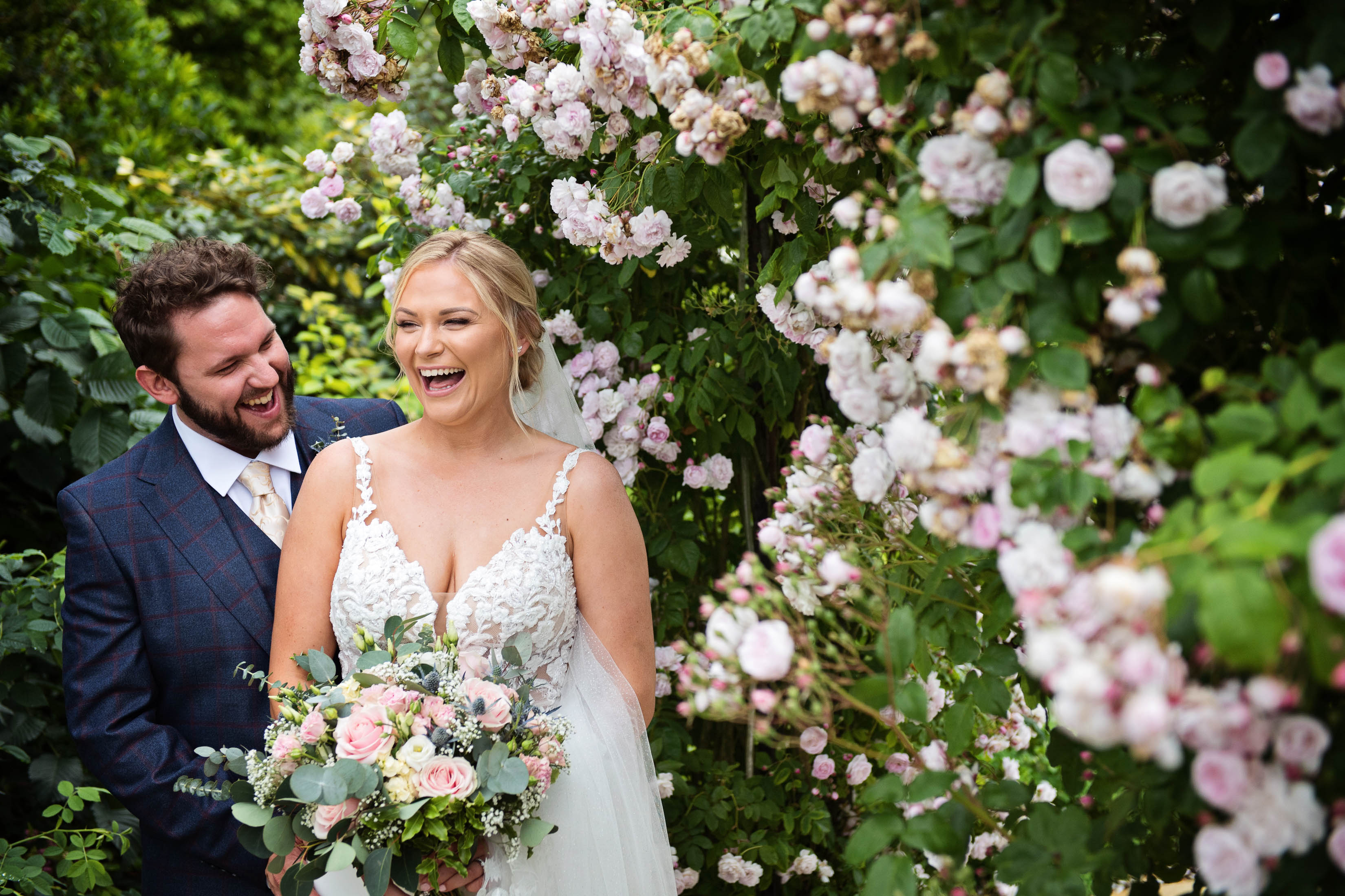 Bride and groom, laughing rather than posing (!), with a gorgeous rose arch to the right of them. 