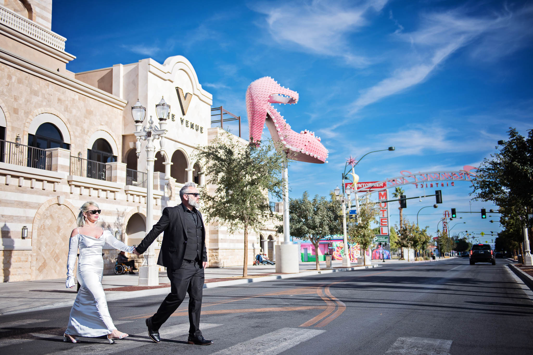 Bride and groom hold hands whilst crossing a Las Vegas street. A large pink stiletto across the street is behind them!   