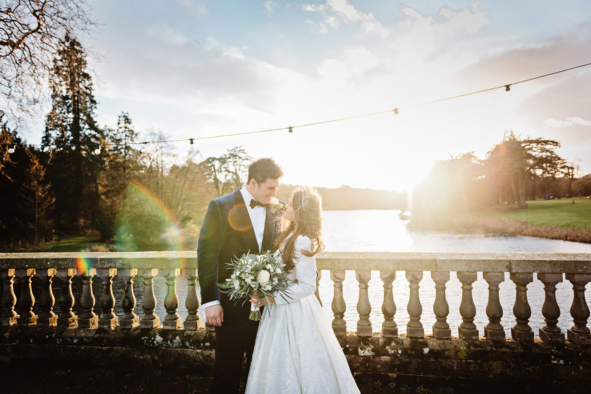 Bride and groom kiss on a bridge over a lake with a stunning sunset in the background