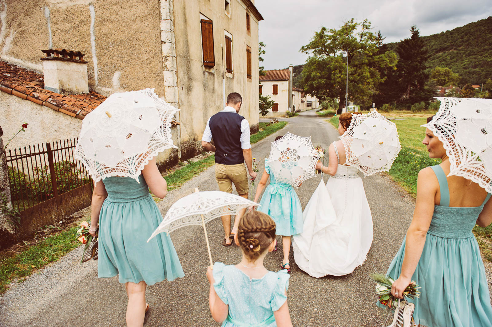 A bridal party walk down a french street, holding umbrella's, photograph behind them. 