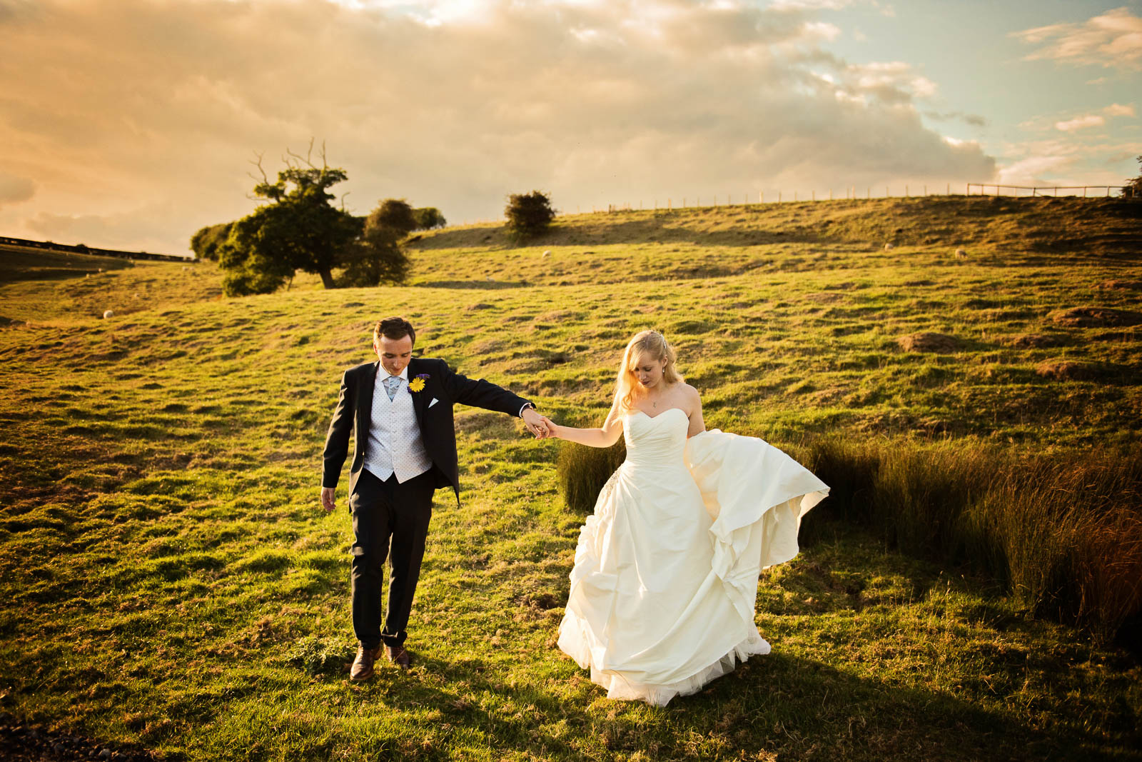 A bride and groom hold hands as they walk across fields with the sun behind them. 