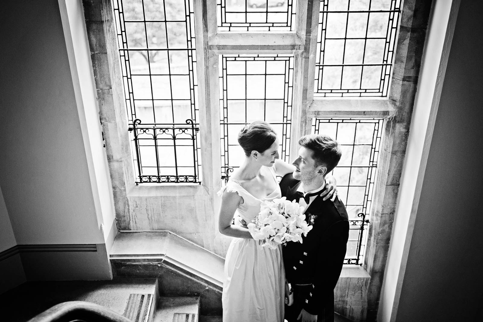 Black & white photograph of Bride & Groom standing (facing each other), with a stain glass window behind them.  
