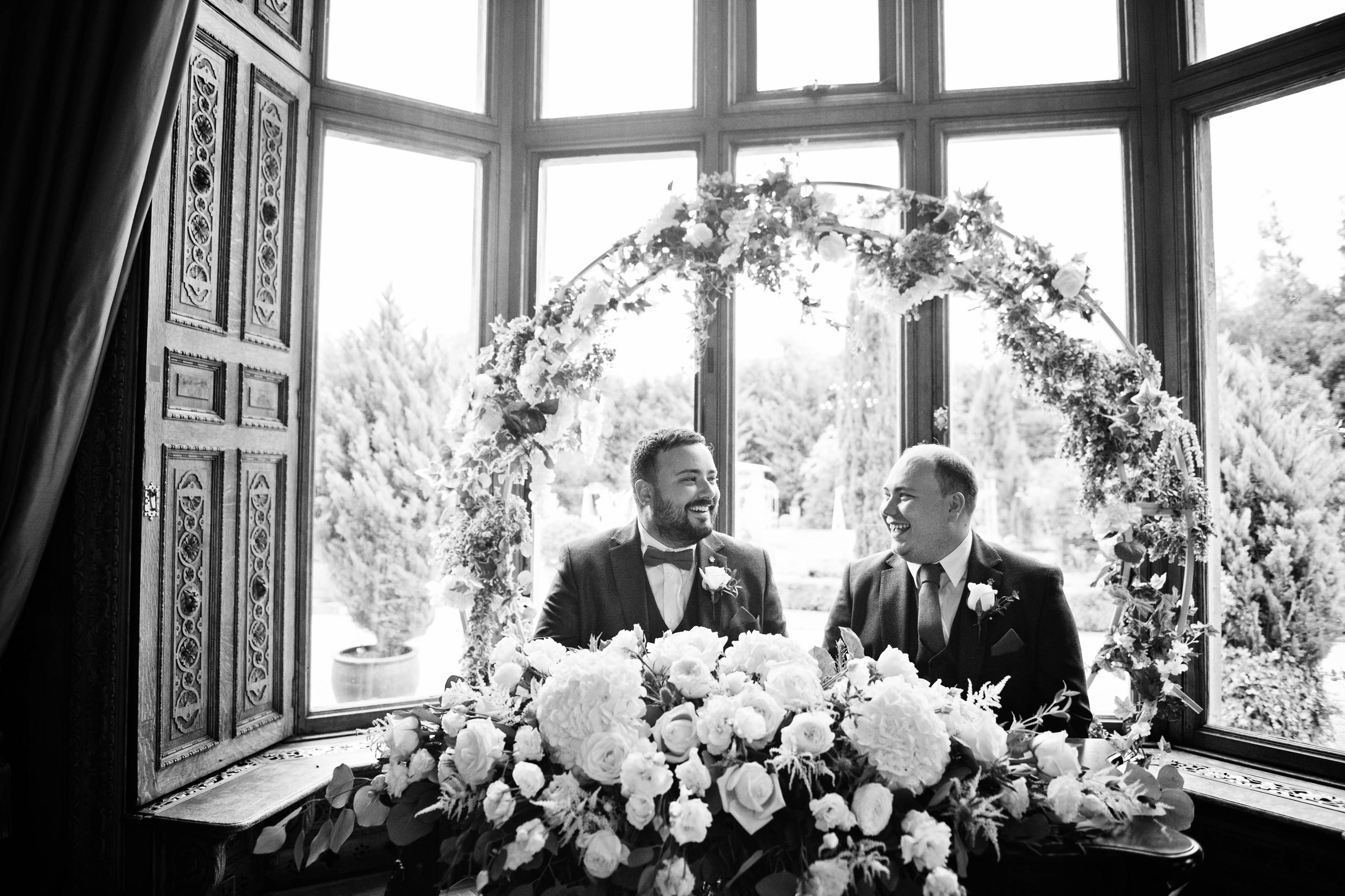 B & W image of husband and husband, siting down, with a bay window and gorgeous flower arch behind them, proudly smiling at each other after signing their wedding certificate, 