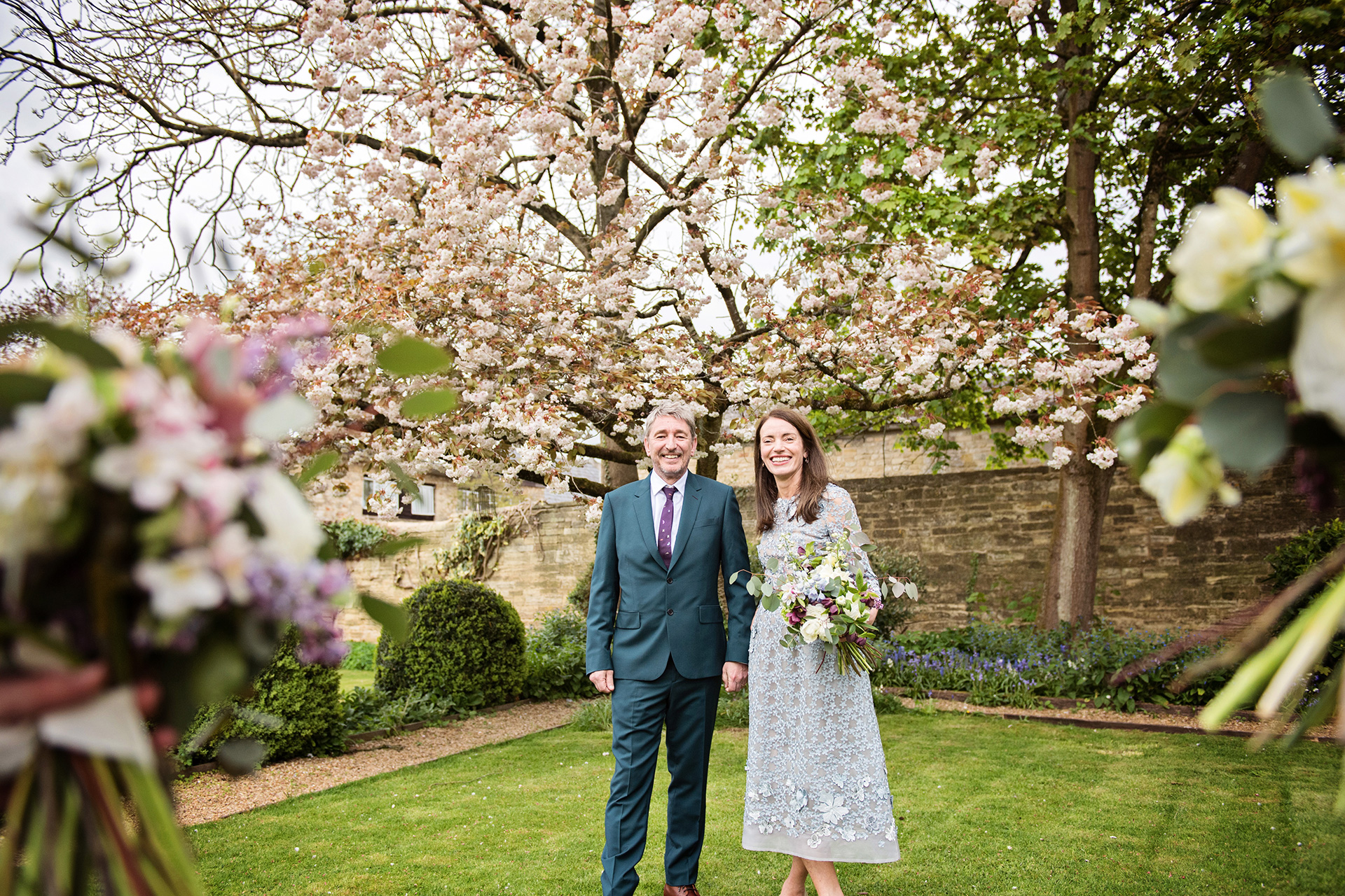 Wedding couple holding hands smiling, with a cherry tree (in blossom) behind them and two bunches of wedding flowers in front of them (framing them). 
