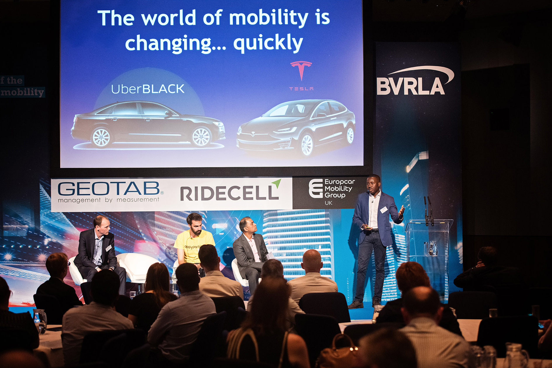 Speaker on stage at a Business conference in the Birmingham ICC with Uber Black and Tesla car.