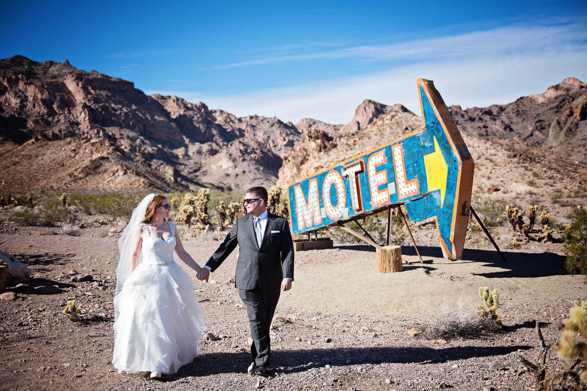 bride and groom hold hands as they walk in the Nevada desert. A lark neon motel arrow sign is behind them.