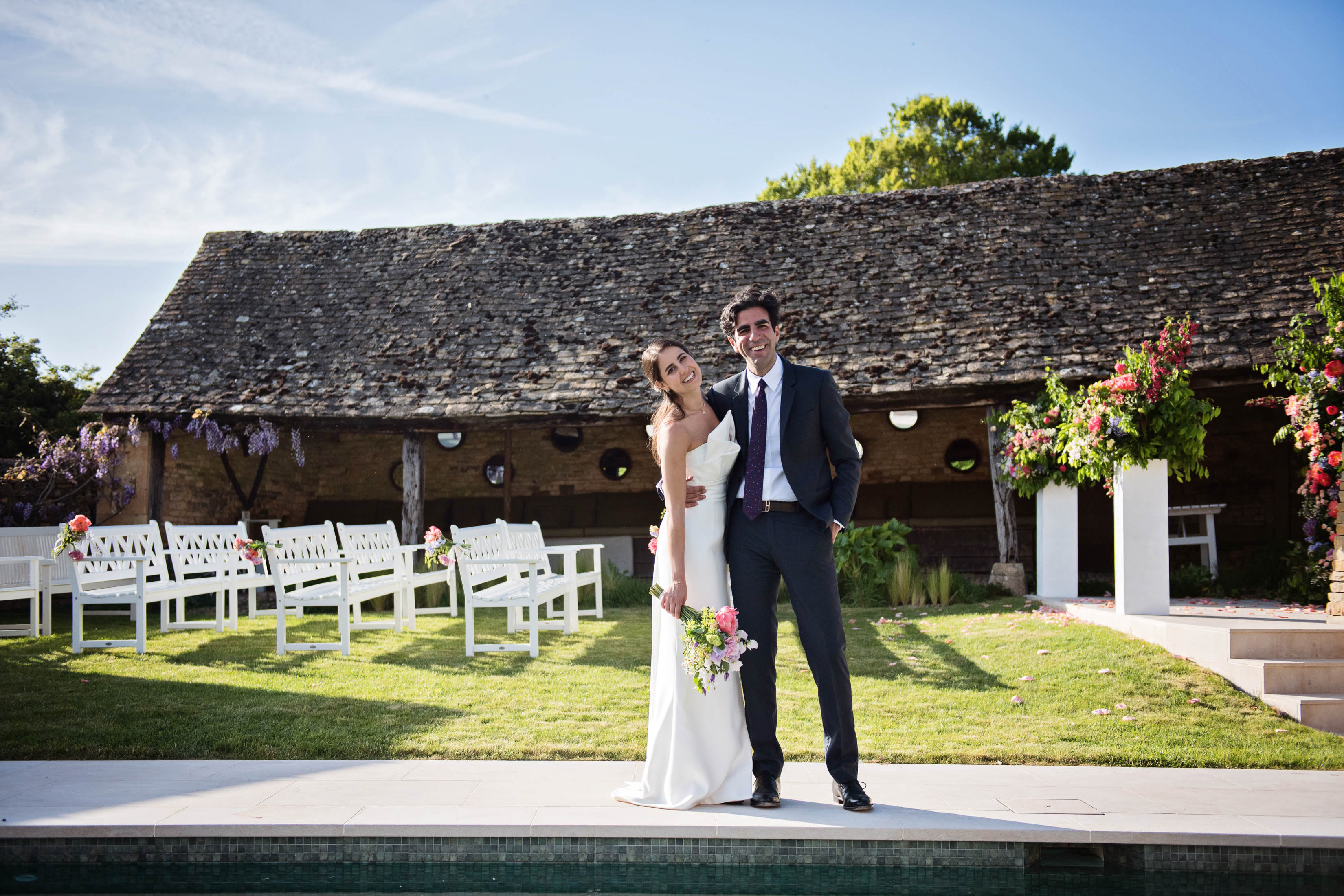 Glamorous Bride & Groom stand in the sunshine together by a swimming pool with their Cotswold wedding ceremony behind them