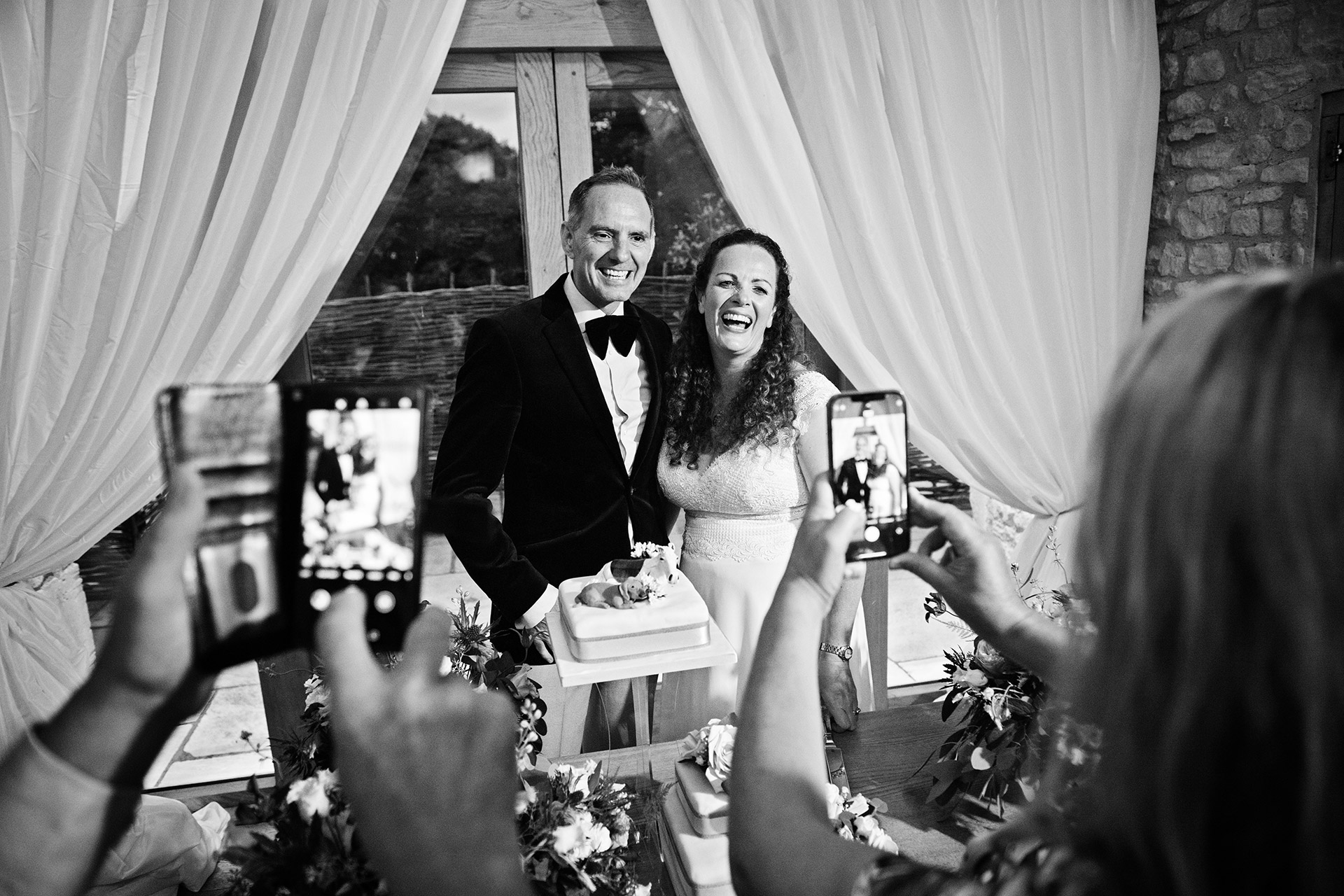 Bride and grooming smiling cutting their wedding cake infront of their friends with with taking photographs with their phones. Black and white image.