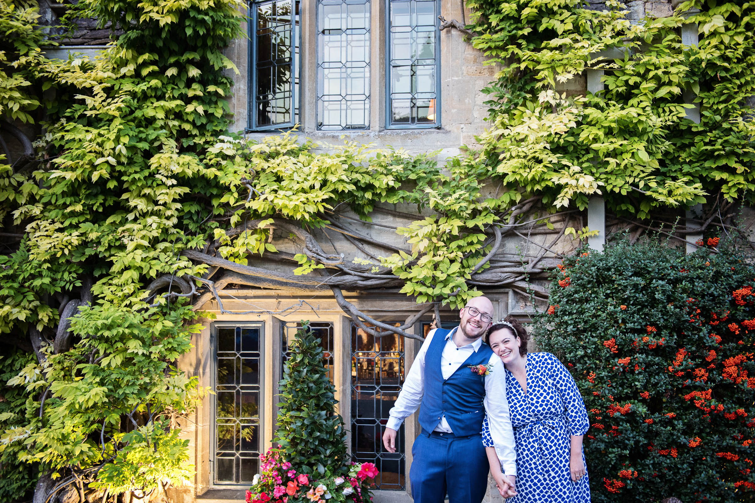Bride & Groom stand shoulder to shoulder, (and hand in hand), smiling with wisteria of The Bay Tree, Burford, behind them.  