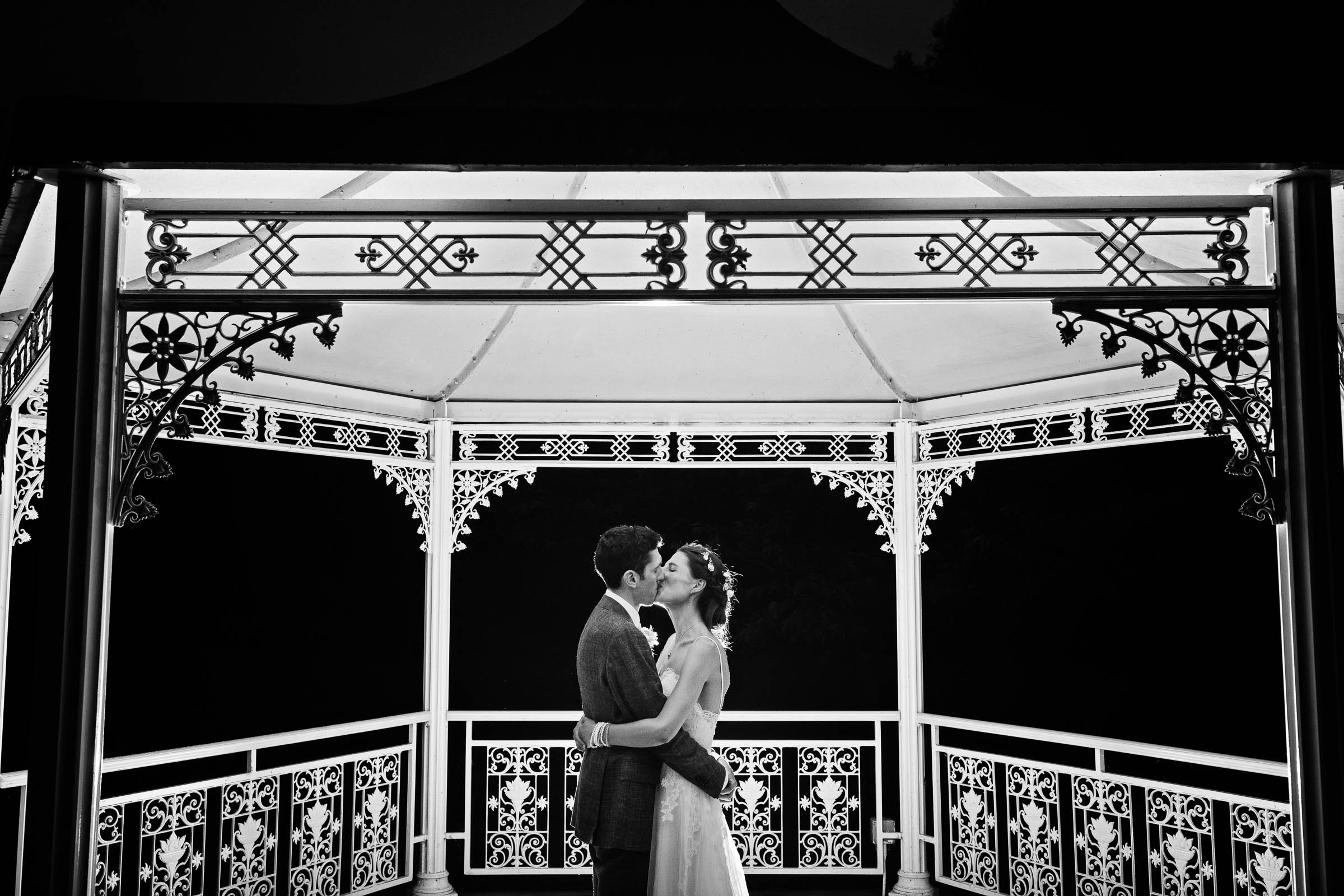 Black and white photograph of bride and groom embracing (with a kiss) underneath a bandstand that is illuminated at night.