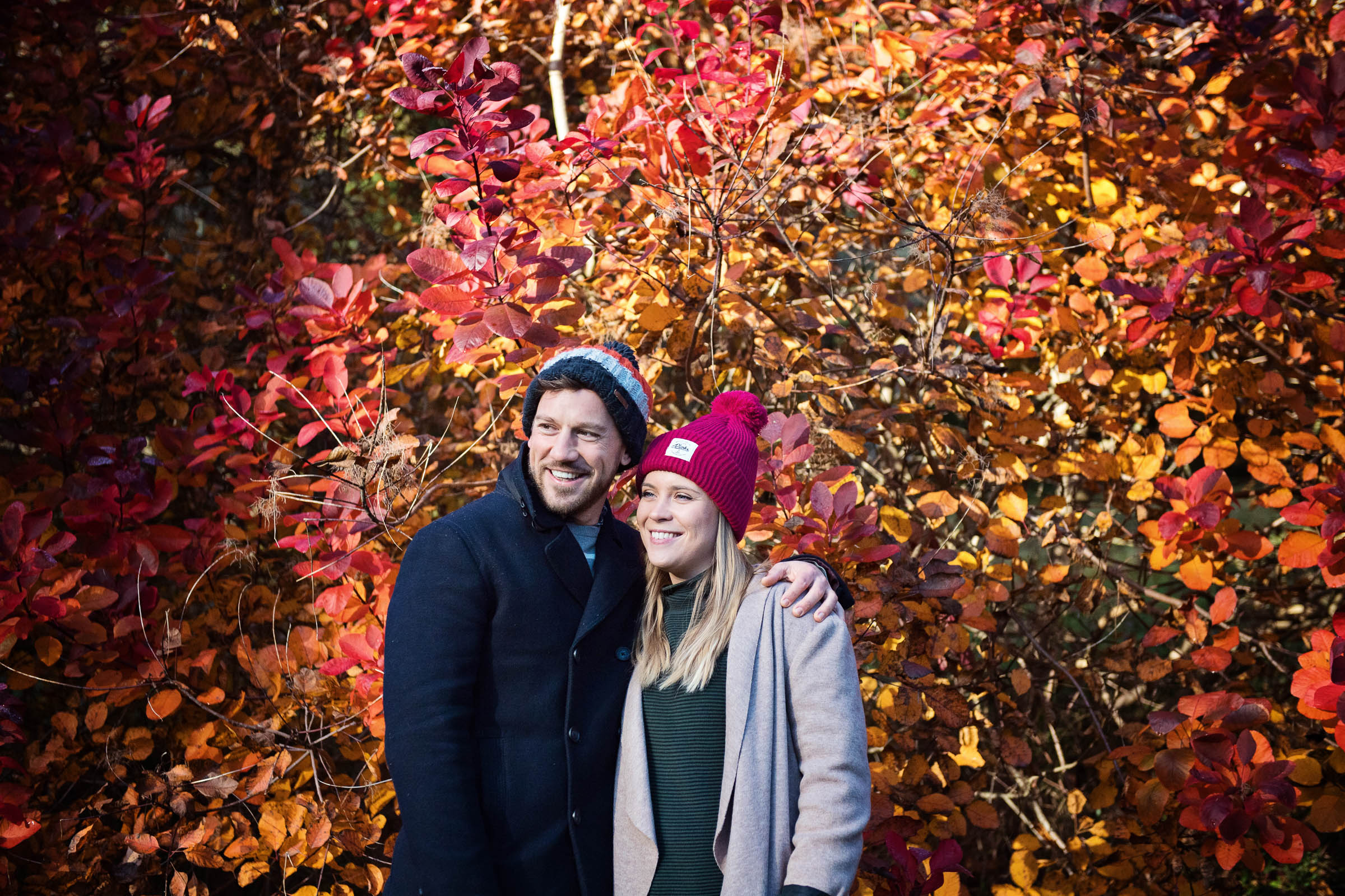 Couple stand smiling (looking away from camera)  with a stunning autumnal red backdrop behind them.