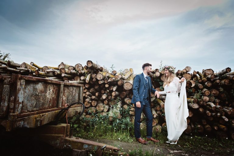 Dramatic photo of bride and groom standing amongst a stack of logs and next to a rusty agriculture trailor. Photo by Blooming Photography