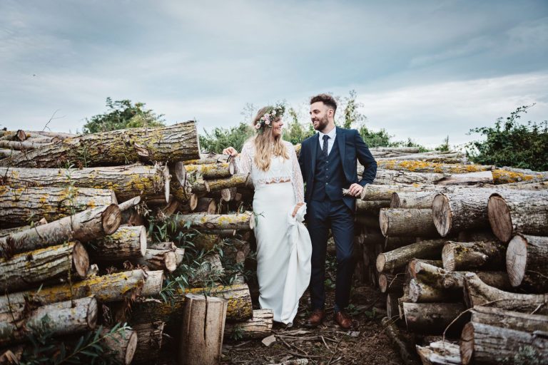 Dramatic photo of bride and groom standing amongst a stack of logs. Photo by Blooming Photography