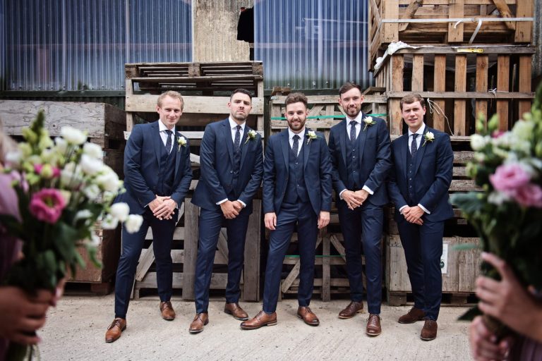 Groomsmen pose up against wooden pallets and surrounded by the bridesmaids flowers.