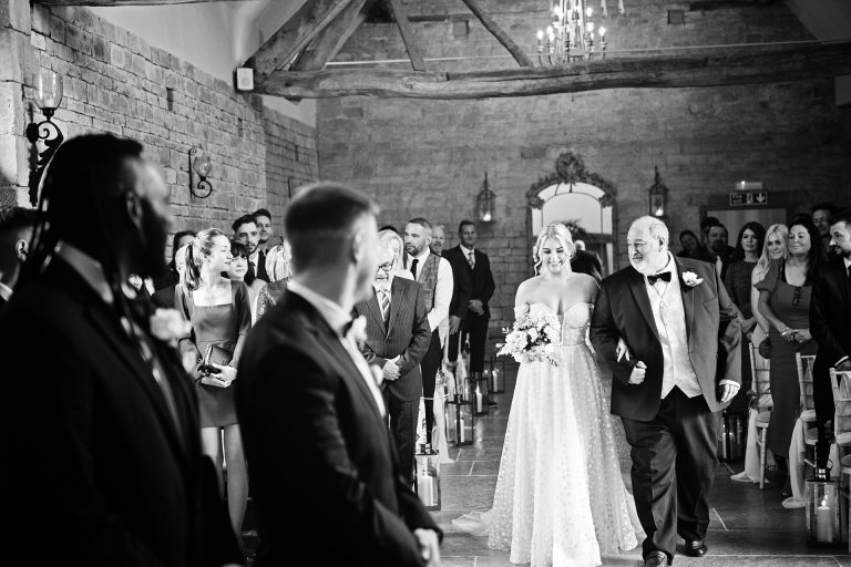 B&w image of the bride being walked down the wedding isle by the father of the bride. The groom and best man look onwards at Blackwell Grange.