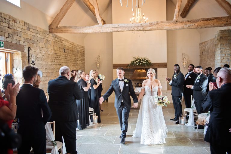 Bride and groom walking down the wedding isle with their guests clapping in celebration. Candid photo by Blooming Photography taken at Blackwell Grange.