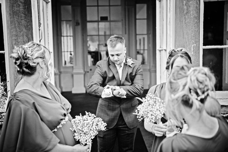 Black and white candid photo of a groomsman looking at his watch with bridesmaids either side of him.