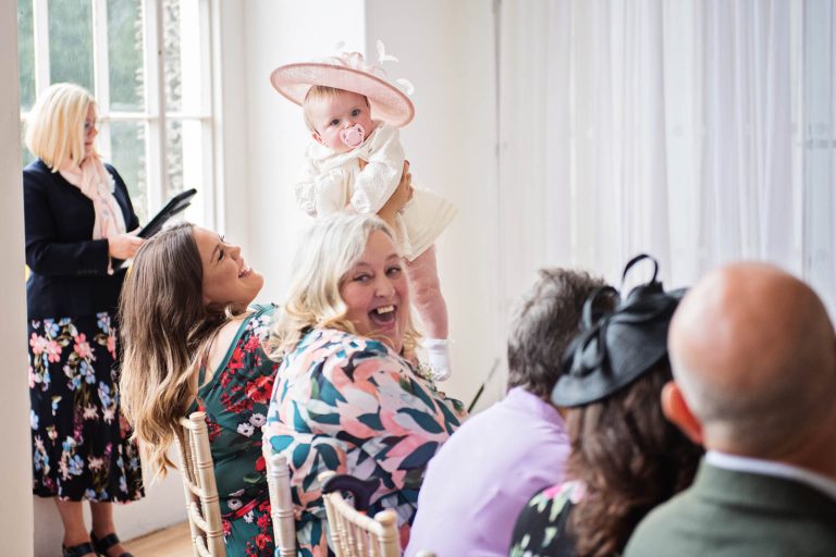 Natural pic of baby wearing a wedding fascinator being held up in the air.