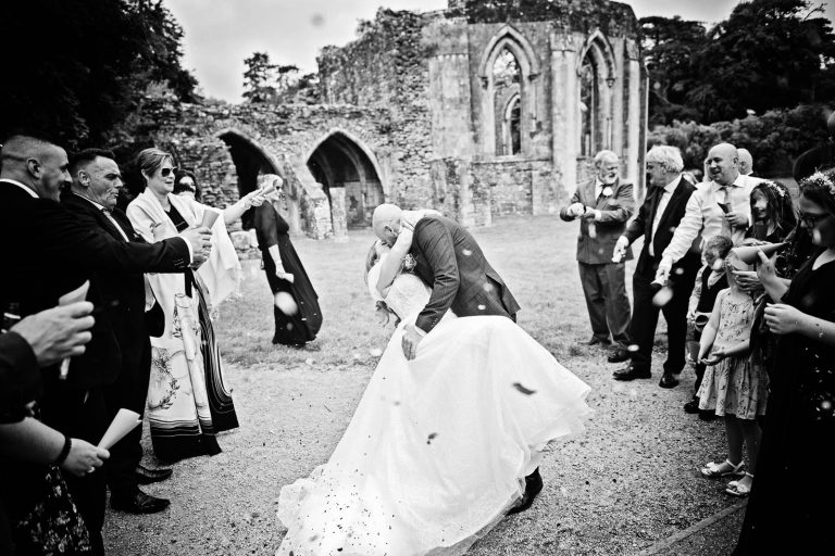 Black & white photo of a bride and groom doing a wedding dip whilst having wedding confetti thrown at them.