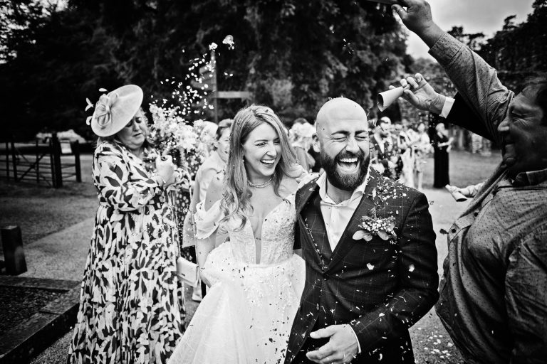 Black & white photo of a bride and groom smiling whilst having wedding confetti thrown at them.