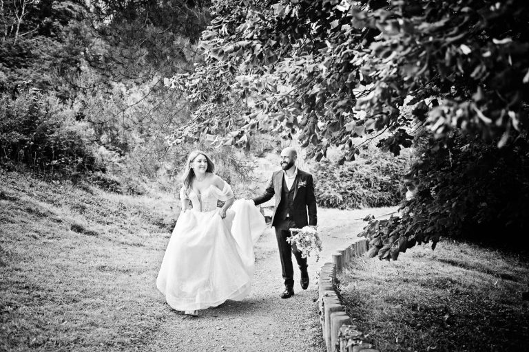 Natural black & white wedding photo of bride and groom walking up smiling whilst the groom is holding the brides dress up.