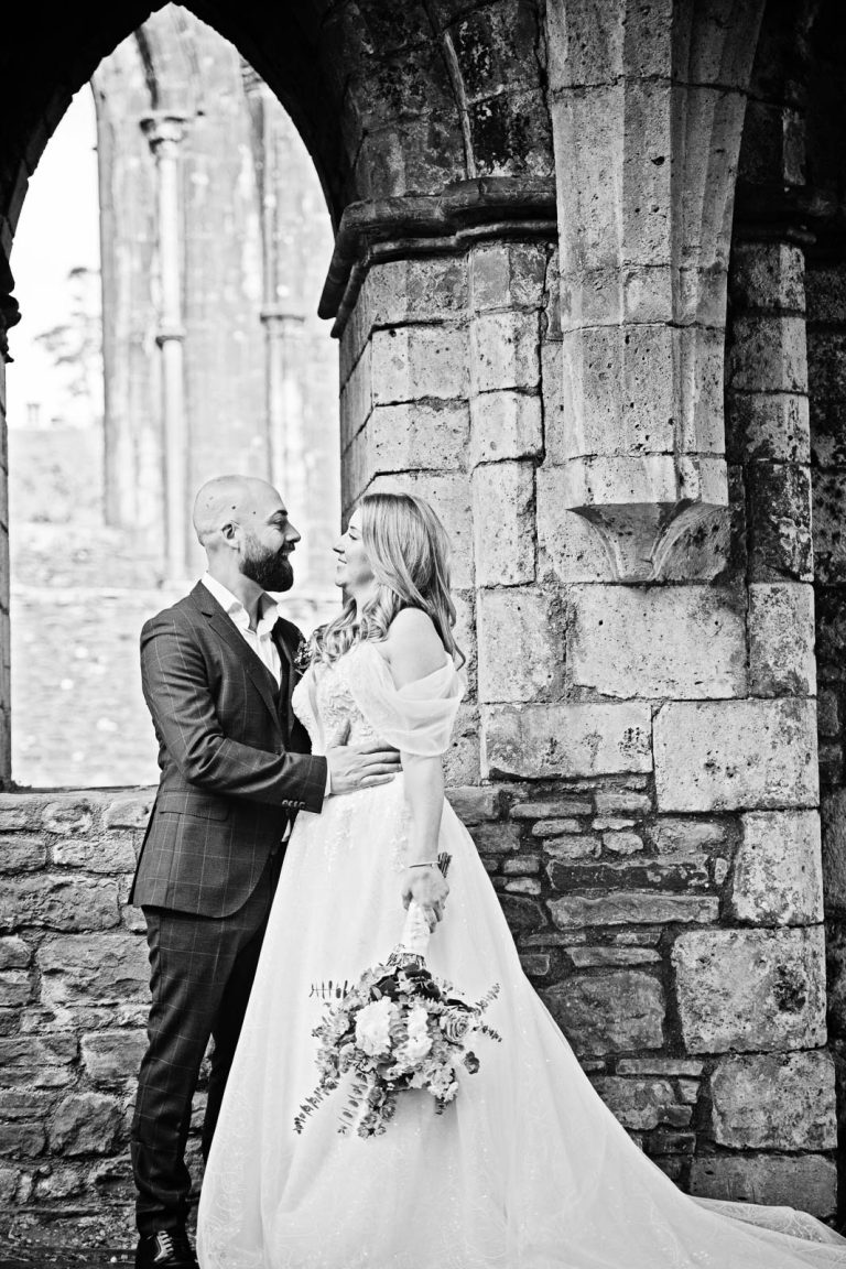 B& W wedding photo of bride and groom holding each other closely, smiling within the ruins at Margam behind them.
