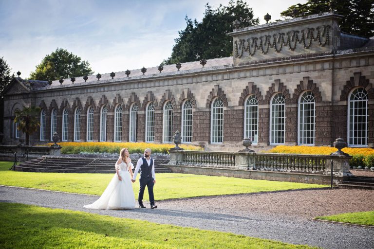 Elegant wedding photograph of bride and groom holding hands and walking down a path with the Orangery behind them.