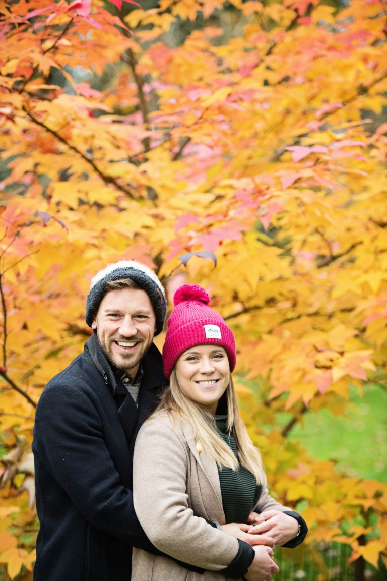 Colourful autumnal backdrop with couple posing in the foreground at Westonbirt Arboretum for their engagement shoot.