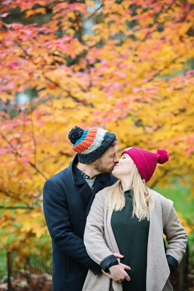 Colourful autumnal backdrop with couple kissing in the foreground at Westonbirt Arboretum for their engagement shoot.