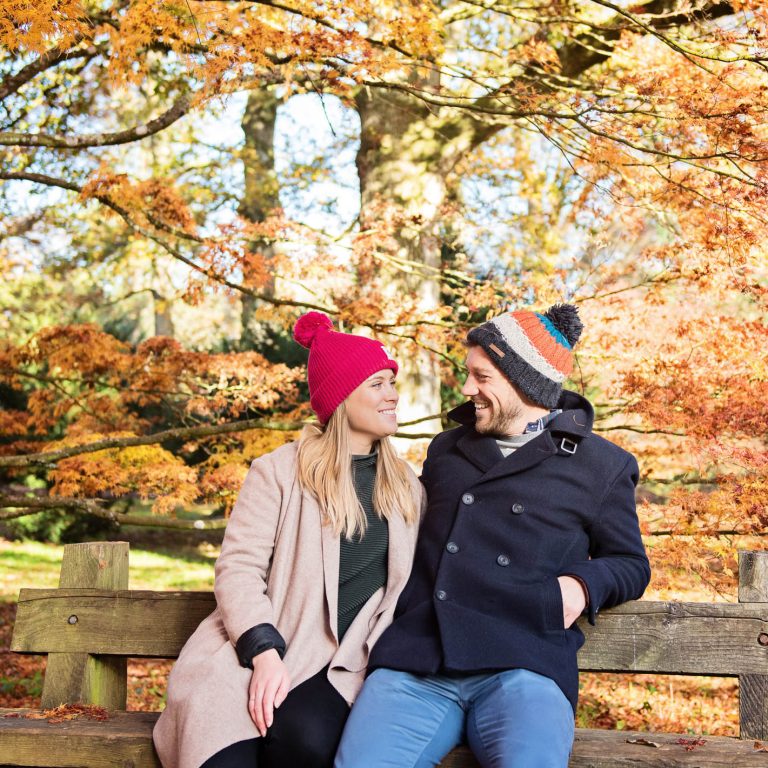 Colourful autumnal backdrop with couple smiling on a wooden bench at Westonbirt Arboretum for their engagement shoot.