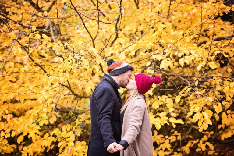 Colourful autumnal backdrop with couple kissing in the foreground at Westonbirt Arboretum for their engagement shoot.