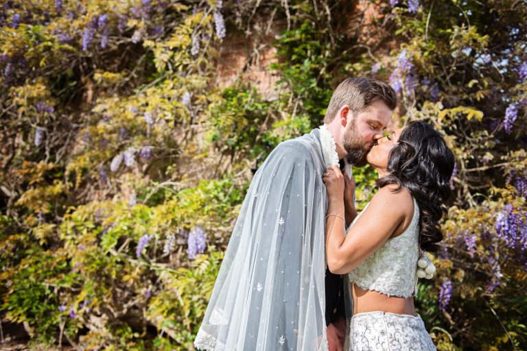 A bride and groom kiss. The groom is wearing the brides' veil around is shoulders, the bride is pulling on the veil ensuring that the kiss is passionate. Wisteria flowers adorn the wall behind them. Photographed at North Mymms Park, by Blooming Photography.