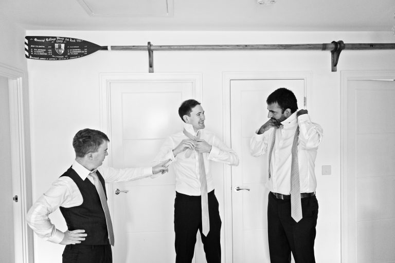 Candid black and white photo of three groomsmen tying their ties, with a Queens College Rowing oar above them.