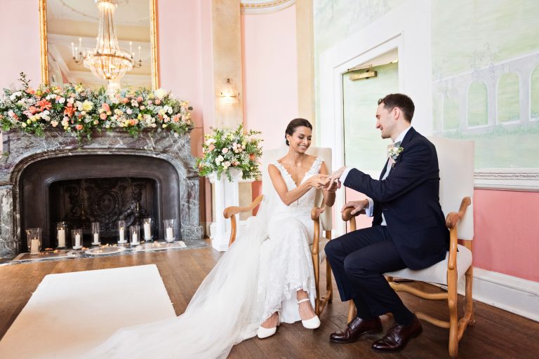 Bride and groom exchange rings whilst sitting down in stately chairs. A gorgeous fireplace with flowers and candles and a mirror reflecting a chandelier adds to the quality of the image.
