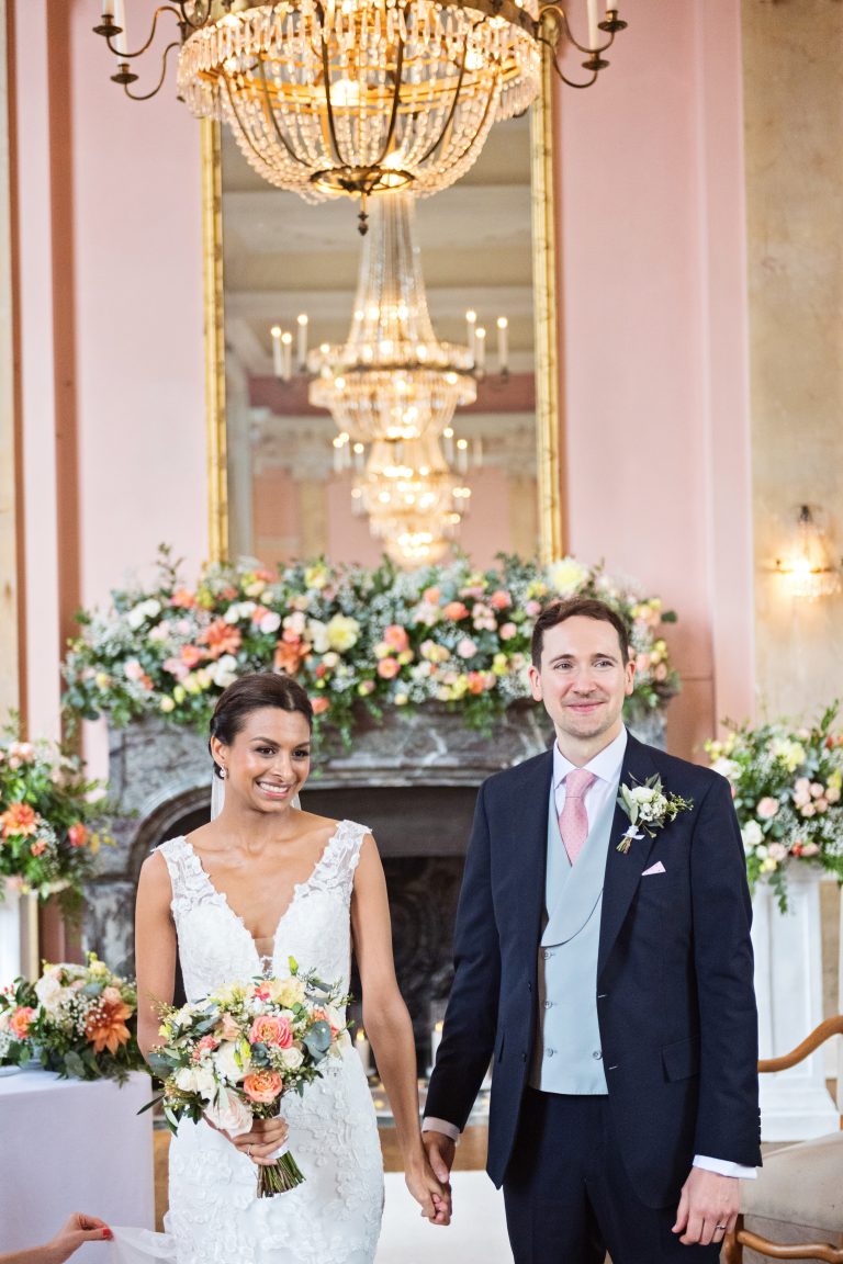 An elegant portrait image of a bride and groom holding hands with a row of chandeliers reflective in the mirror. Photography by Blooming Photography at Danesfield House Hotel and Spa.