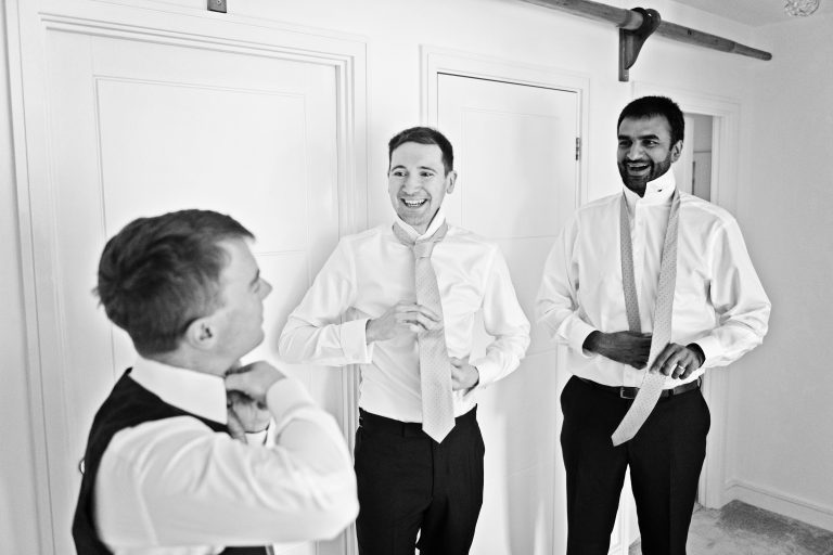 Candid black and white photo of three groomsmen tying their ties smiling.