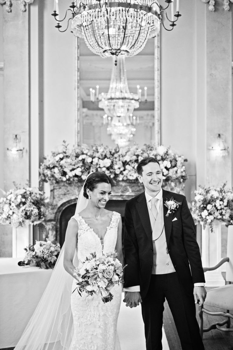 An elegant portrait image of a bride and groom holding hands and smiling with a row of chandeliers reflective in the mirror. Black and white image.