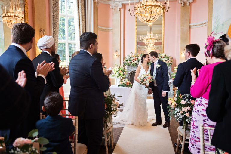 Wedding guests applaud the bride and groom who seal their marriage with a kiss in the grand hall at Danesfield House Hotel.