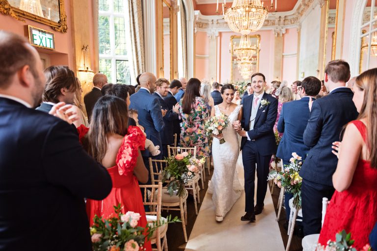 A bride and groom walk down the wedding isle together at Danesfield House Hotel and Spa with their wedding guests clapping. Elegant image.
