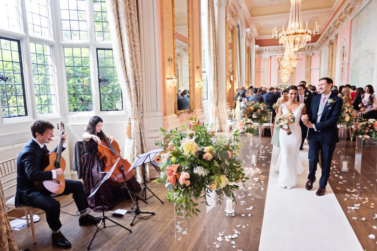 A bride and groom walk down the wedding isle together but pause at seeing the musicians play a beautiful classical piece of music at Danesfield House Hotel and Spa. Elegant image.