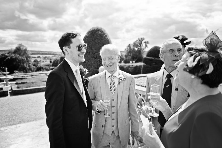 The groom chats to his parents informally, with the mother telling stories whilst the father is smiling in the gardens of Danesfield House Hotel and Spa. Black and white photo