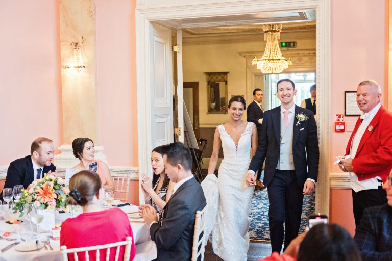 Bride and groom enter the wedding breakfast room at Danesfield House Hotel and Spa smiling with the friends and family smiling and clapping and taking photographs with their mobile phones.