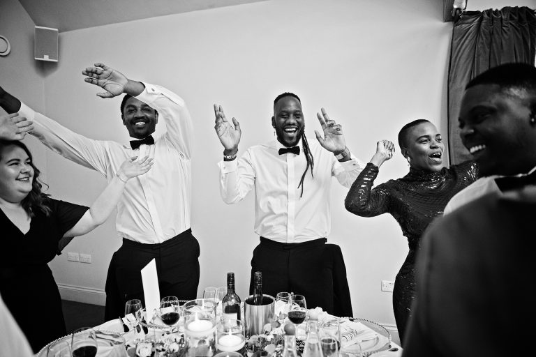 B & w image of wedding guests smiling and singing with their hands in the air during a song. Party! Candid photo by Blooming Photography. Taken at Blackwell Grange, Warwickshire.