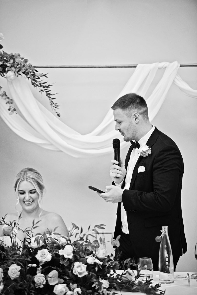 B&W image of the Groom delivering his speech with the bride smiling next to him. Candid photo by Blooming Photography. Taken at Blackwell Grange, Warwickshire.