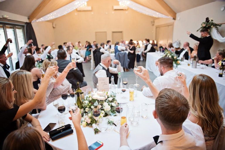 Wedding guests raise their glasses to say cheers at the end of the speeches. Candid photo by Blooming Photography. Taken at Blackwell Grange, Warwickshire.